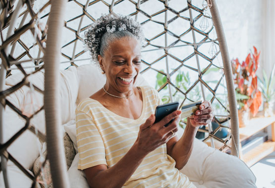 Women holds a credit card and smiles as she looks at her smart phone. She is sitting in a hanging wicker swing.